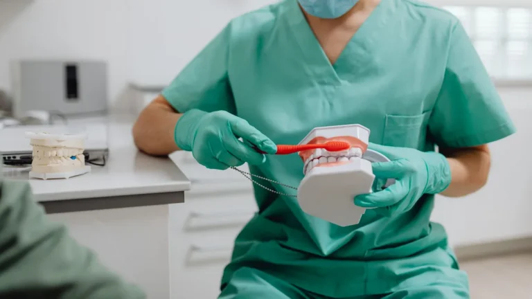 Dental professional in scrubs demonstrates proper brushing on a model in a clinic setting.